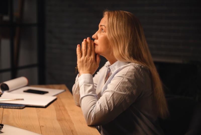 older adult female praying seated at a table in a dark room
